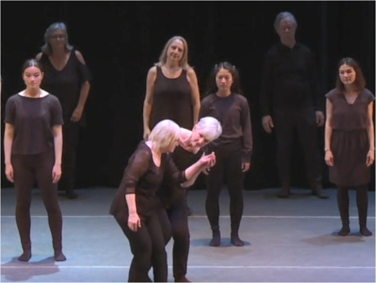 people on stage wearing black watching a center duet.. one white haired woman holding hand of another white haired woman